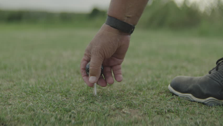 Close-up of a male hand placing a black golf ball onto a tee on a golf course, focused pre-shot moment, detailed sports scene, manicured grass, natural outdoor light, calm and cinematic golf atmosphere.