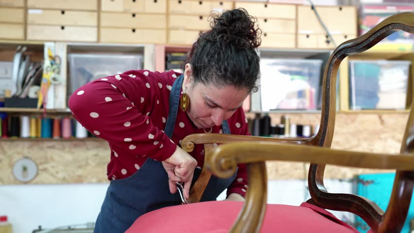 Middle aged woman in her workshop meticulously restoring antique furniture. She is carefully cutting and fitting a new red fabric cover