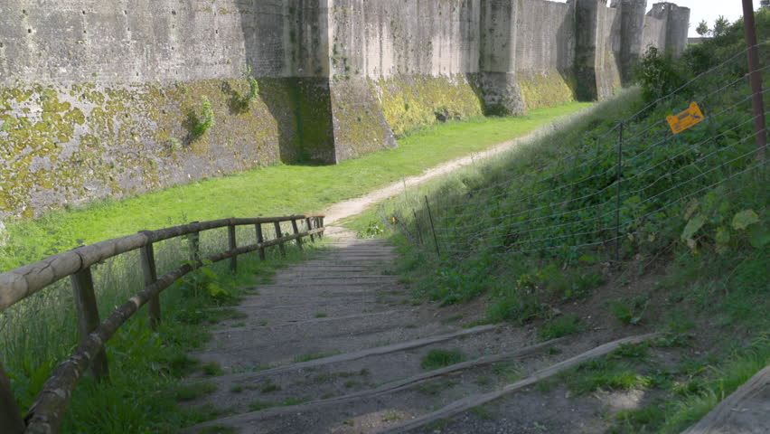 Ancient stone fortress wall with defensive towers and a grassy moat on a bright sunny day.