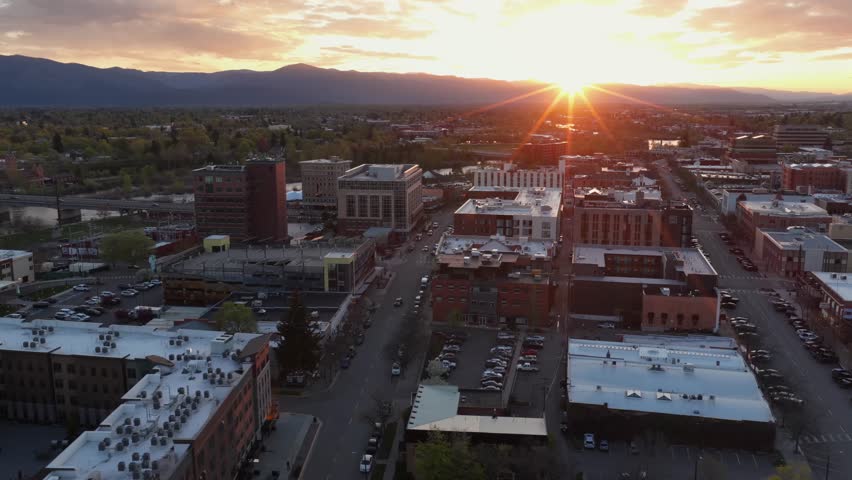 Missoula downtown red brick buildings and flat rooftops glowing under intense golden sunburst sunset, parking lots and streets stretching to mountain range silhouette. Montana, USA. Aerial rising wide