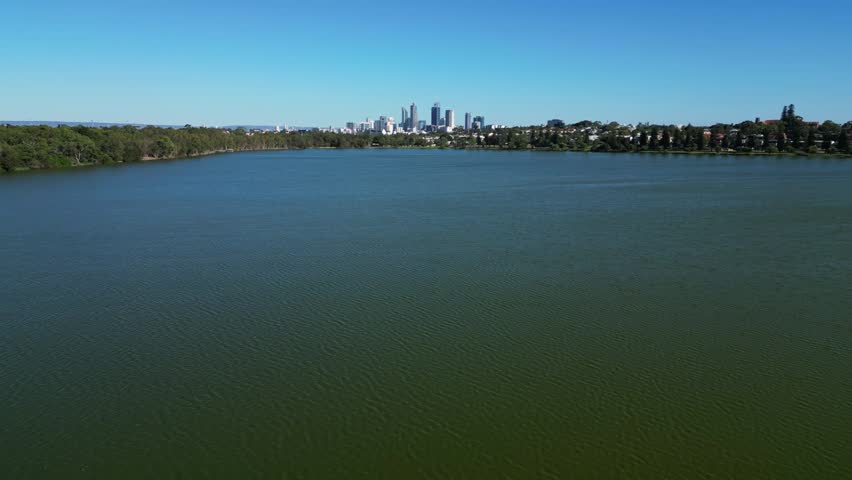 Forward flight over vast calm urban lake with tree lined shores and full Perth city skyline rising above the horizon under clear blue sky. Galup Reserve, Perth, Australia