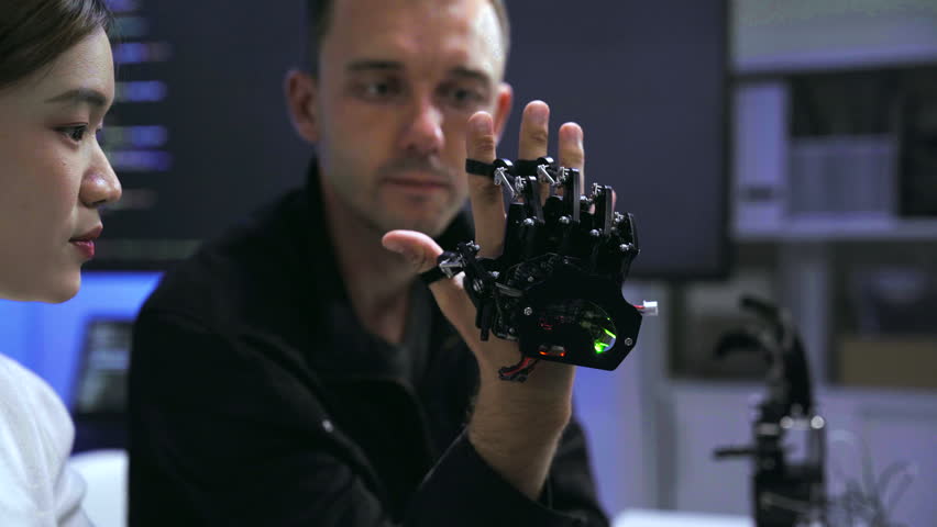 Young Asian woman and Caucasian man collaborating on robotic hardware device. Tech developers examining electronic components in modern laboratory with blue ambient lighting.