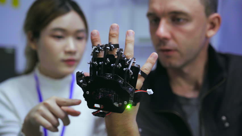 Young Asian woman and Caucasian man collaborating on robotic hardware device. Tech developers examining electronic components in modern laboratory with blue ambient lighting.