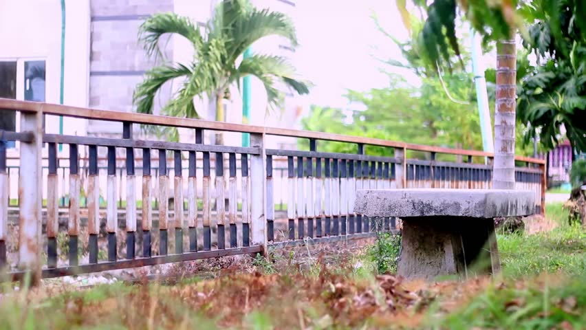A concrete stone bench in a grassy park area next to an old iron fence with a blurry palm tree background.