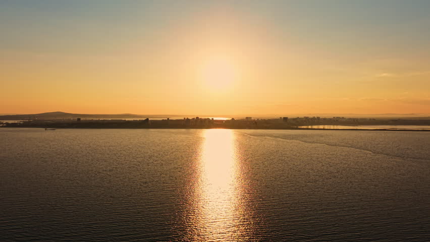 A sunset creates a bright scene above the water with sunlight reflecting on the surface. The city skyline is visible in the distance as evening begins.