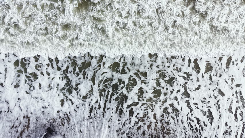 Waves roll onto a beach, breaking against the dark sand and rocky shore. White foam forms as the water recedes. The scene shows the shoreline under bright daylight.