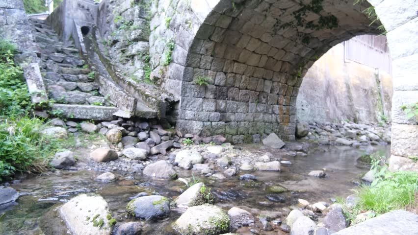 Silent small stream flowing under old stone bridge with rocks and greenery