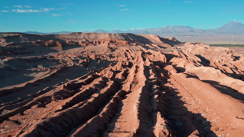 Backward flight over dramatic eroded salt rock ridges and sand dunes revealing San Pedro de Atacama oasis and snow-capped volcanic peaks on the horizon. Valle de la Luna, Chile