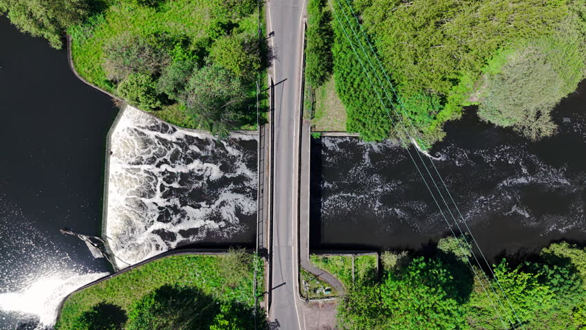 Aerial view of a river weir with cascading water and a bridge surrounded by lush green trees in Kingsbury, England, United Kingdom.