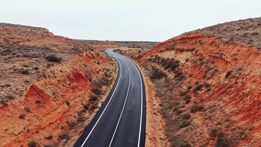 A road winds through a landscape of red hills. The scene shows an empty highway with no cars. The sky is overcast, creating a gray setting. The hills are bare with some shrubs.