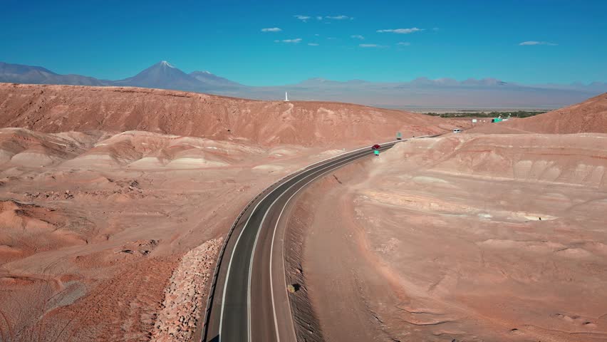 Ascending aerial over winding highway through red desert badlands with vehicles heading toward San Pedro de Atacama oasis and iconic volcanic peak on the horizon. San Pedro de Atacama, Chile