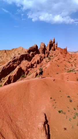 Aerial view of Skazka Canyon with a man standing on a ridge overlooking red sandstone formations under a blue sky in Tosor, Issyk-Kul Region, Kyrgyzstan.