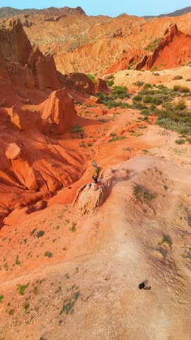 Aerial view of a person standing on a ridge among the red sandstone formations of Skazka Canyon under bright sunlight in Tosor, Issyk-Kul Region, Kyrgyzstan.