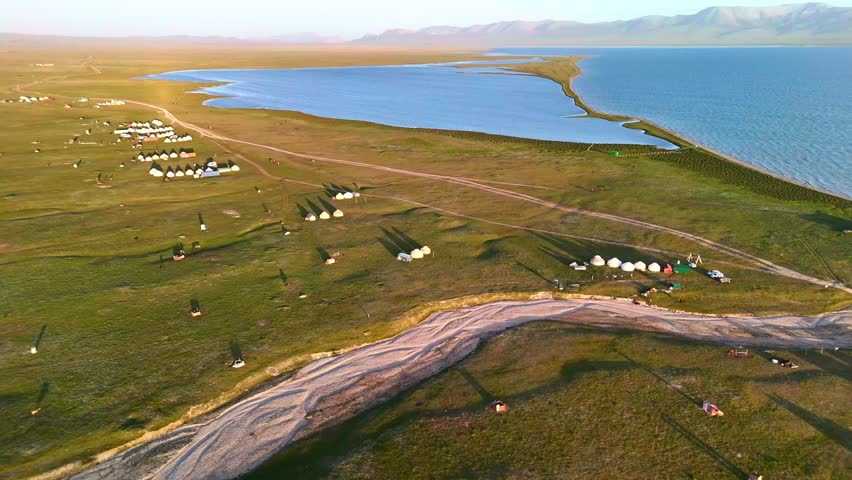 Aerial view of Song-Kul lake with traditional yurt camps scattered across the green plains under golden hour light in Naryn Region, Kyrgyzstan.