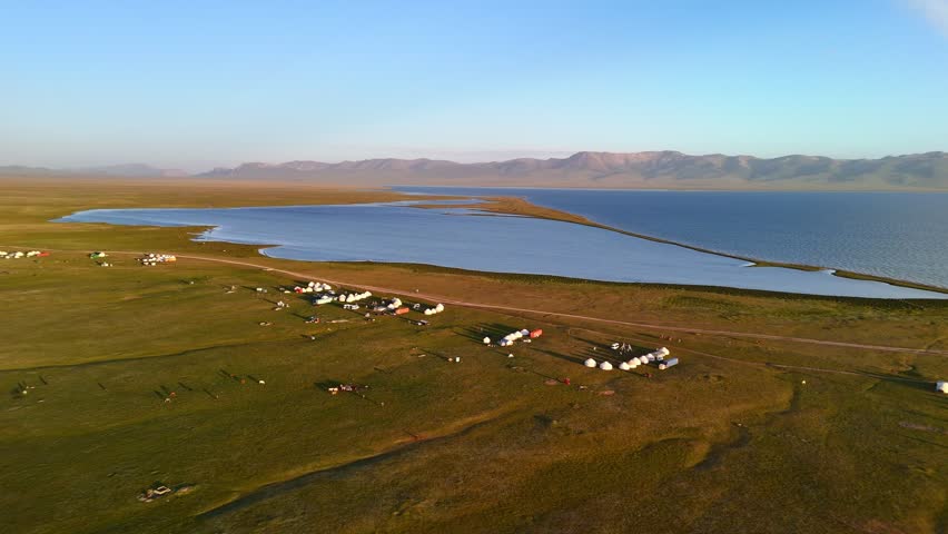 Aerial view of Song-Kul lake with traditional yurt camps scattered across the green alpine pastures under a clear sky in Naryn Region, Kyrgyzstan.