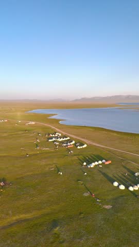 Aerial view of traditional yurt camps on the green shores of Song-Kul lake under a clear sky in Naryn Region, Kyrgyzstan.