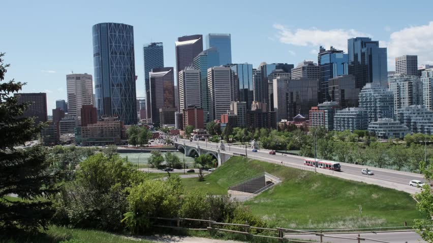 Calgary downtown skyline footage with a river in the foreground on a sunny day. Traffic moves on a road with bridges, green trees, and grass lining the riverbanks. The cityscape features modern buildings and condominiums under blue sky.