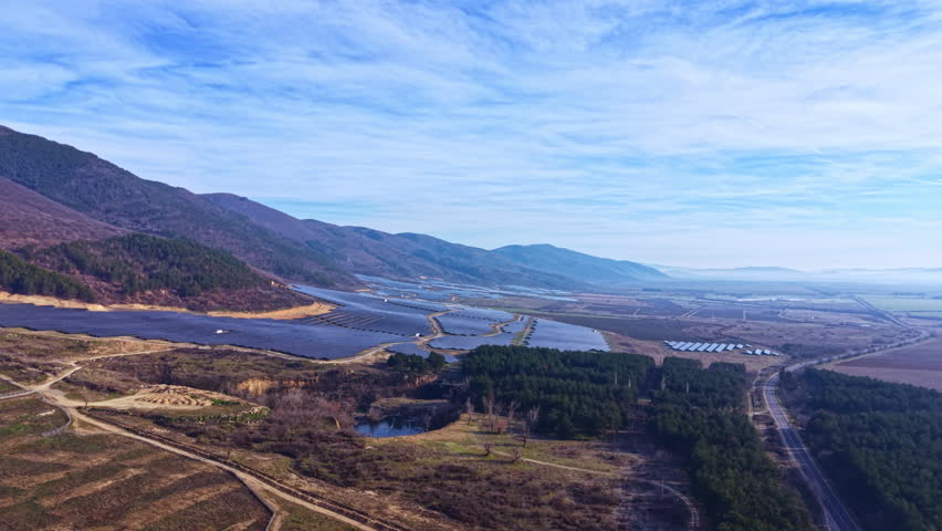 Fields and solar panels stretch across the land near mountains with light clouds in the blue sky above. The scene shows water bodies and natural landscape under sunlight.