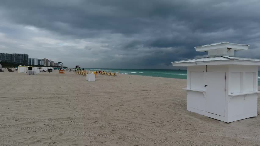 Aerial view of South Beach featuring a white kiosk on the sand under a dark stormy sky with turquoise water and distant city skyline in Miami Beach, Florida, United States.