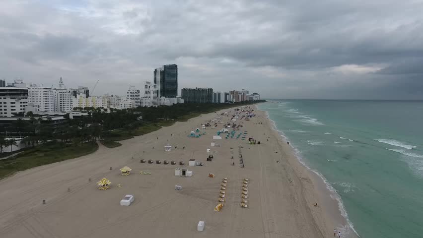 Aerial view of South Beach featuring the sandy coastline, turquoise ocean waves, and high-rise buildings under a cloudy sky in Miami Beach, Florida, United States.