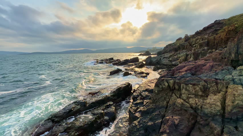 Waves hit the rocky shore as the sun begins to set behind clouds. The scene shows dark rocks and water reflecting light in a coastal area during evening.