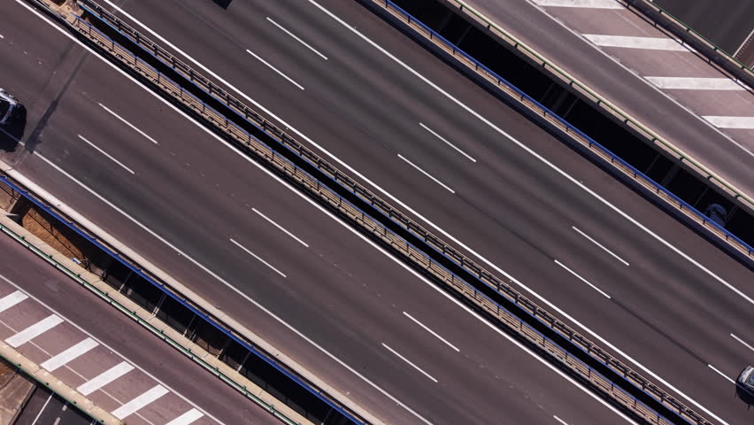 Top-down overhead view of a multi-lane highway interchange with cars and trucks in motion, featuring road markings, barriers, and surrounding vegetation