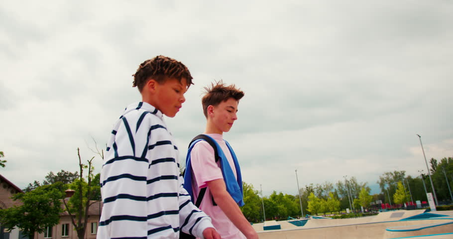Two multiracial teenage boys walking and talking in a skatepark. One friend is bouncing a basketball while they enjoy their leisure time