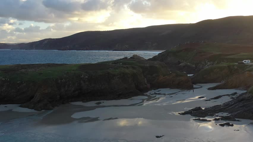 Aerial view of Ermita de Santa Comba on a rocky island surrounded by rugged cliffs and a sandy beach during golden hour in Covas, La Coruna, Spain.