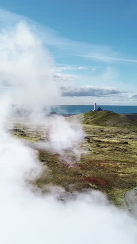 Aerial drone view of steaming geothermal vents across the rugged lava fields of the Reykjanes Peninsula. A winding road leads toward a lighthouse near the Atlantic Ocean. Vertical