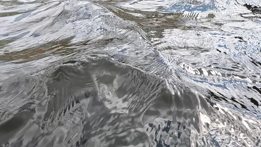a wide surface of river water with a blue sky and reflections of clouds, dark outlines of rocks are visible below them, light ripples moving along small waves in daylight