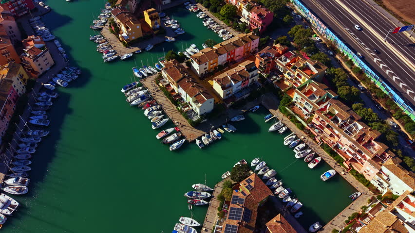 Boats are parked in a channel with green water surrounded by buildings. A busy road runs parallel to the marina. It is daytime with clear sunlight.