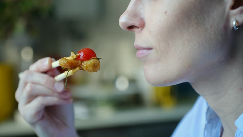 Profile view of young caucasian woman enjoying shrimp, cherry tomato, holding food with wooden chopsticks and putting in mouth for tasty lunch. Female eating shrimp and cherry tomato with chopsticks