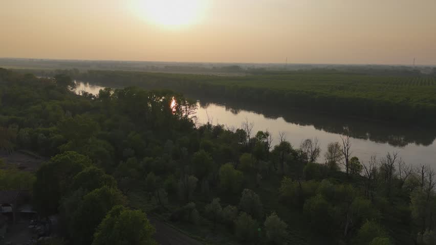 Po River flowing through riparian forest and agricultural lowlands near Isola Serafini in Piacenza province, Emilia Romagna, Italy, during sunset with reflective water, floodplain habitat drone ascent