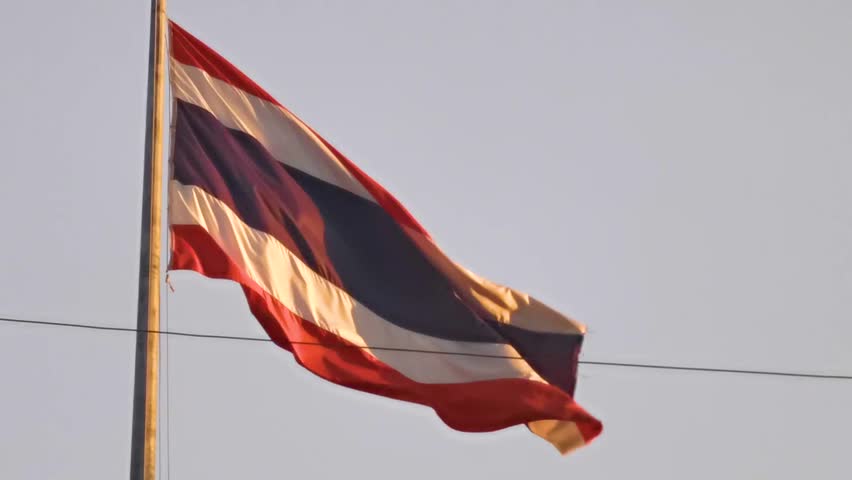 Thai flag waving at sunset with warm golden sky and tropical atmosphere creating calm patriotic coastal scene in Southern Thailand in Southeast Asia.