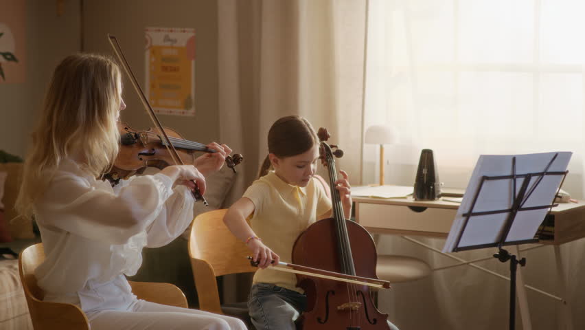 Medium long shot of Caucasian young adult teacher playing violin and instructing female child playing cello during home lesson. Ideal for music school marketing and educational campaigns