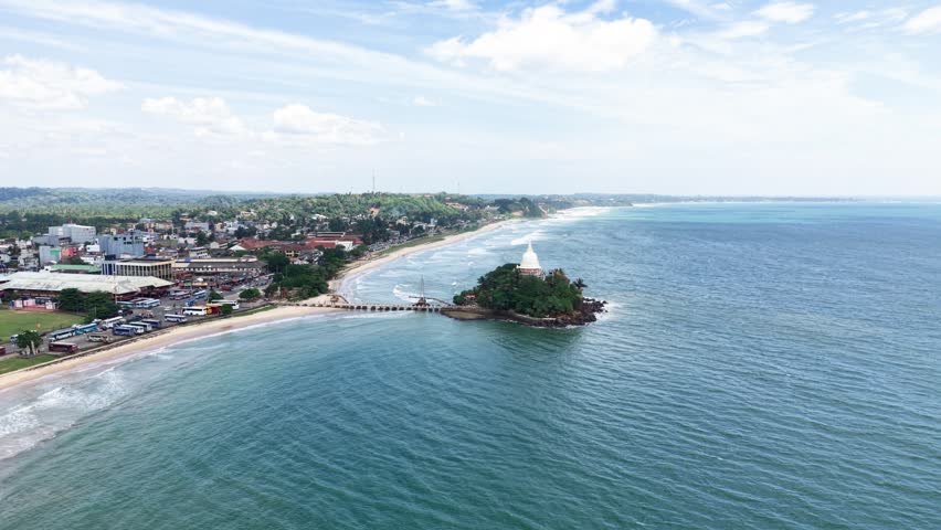 Aerial view of Parewi Duwa Temple on a small island connected by a bridge to the sandy beach and coastal town of Matara, Sri Lanka.