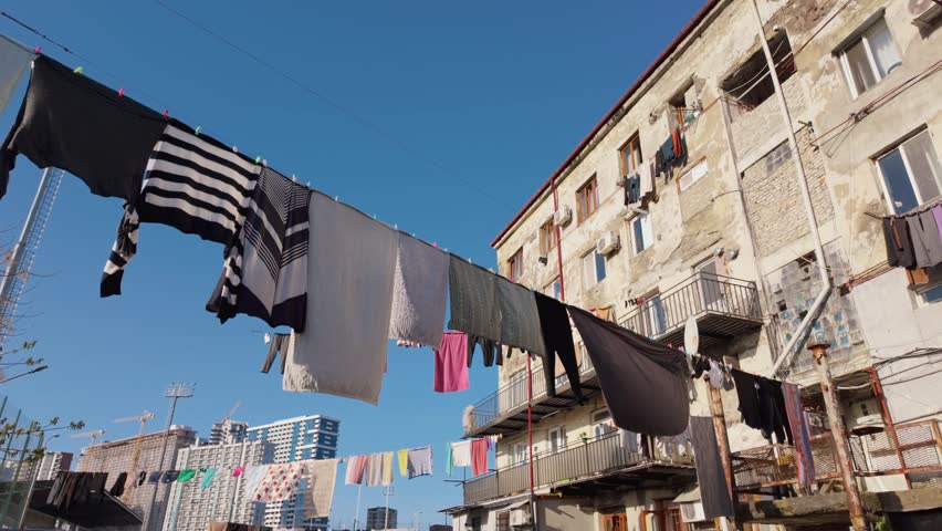 Low-angle view of laundry drying on clotheslines strung between old, weathered apartment buildings in a poor neighborhood, showing a stark contrast with modern skyscrapers in the distance
