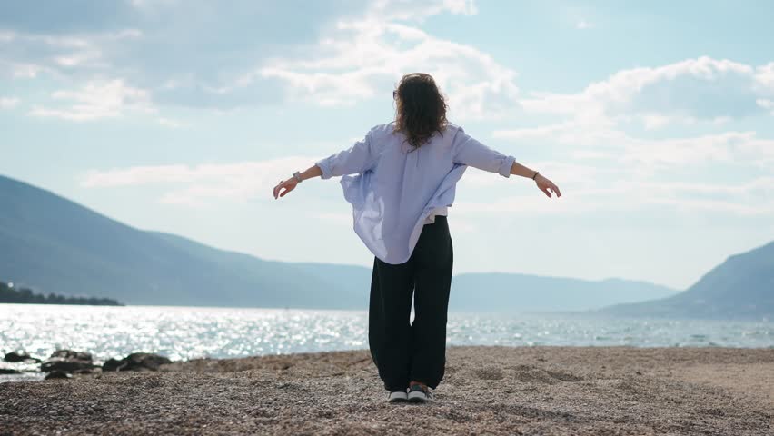Slow motion shot of a young woman standing on the seashore with arms outstretched, while her hair and shirt flow in the wind, expressing freedom and connection with nature