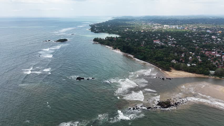 Aerial view of the tropical coastline with waves crashing against rocks and lush green trees along the sandy beach in Matara, Southern Province, Sri Lanka.