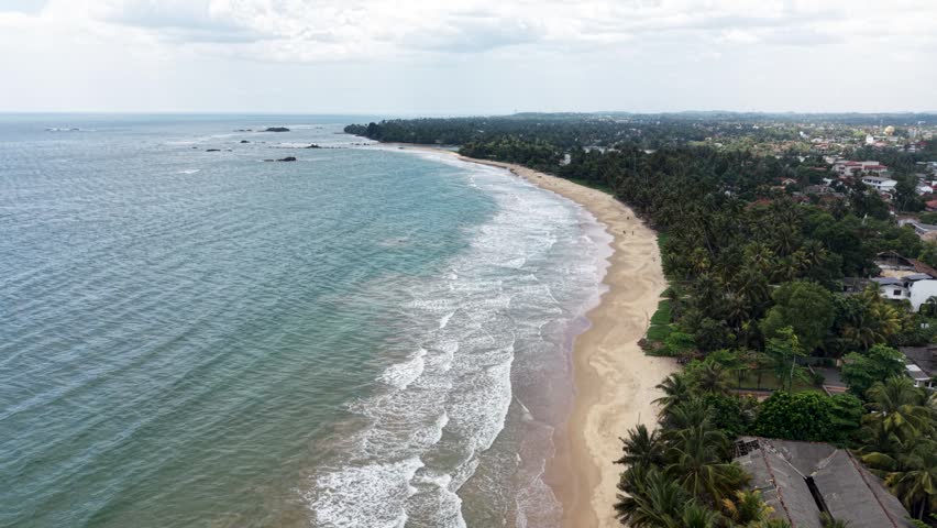 Aerial view of the turquoise ocean waves crashing on a sandy beach lined with palm trees and coastal buildings in Matara, Southern Province, Sri Lanka.