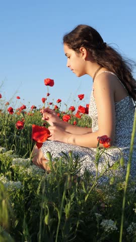 Girl sitting cross-legged in a poppy field smelling a red poppy