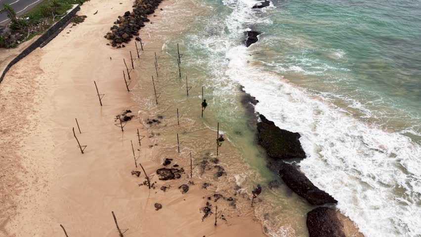 Aerial view of traditional stilt fishing poles with fishermen on a sandy beach with waves crashing against rocks in Koggala, Southern Province, Sri Lanka.