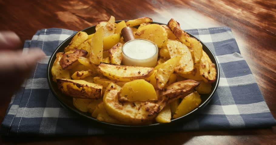 Close up of hands rotating a plate filled with crispy roasted potato wedges. A small cup of creamy dipping sauce is placed in the center for dipping potato wedges before eating.