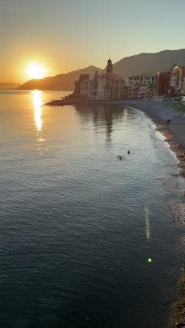 People walk along the beach at sunset in Camogli, Italy. Waves gently lap against the shore as the sun sets behind the buildings. The sky shines with orange light reflecting on the water.