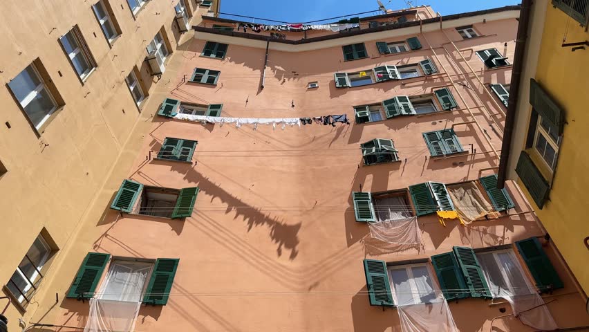 Residents hang clothes on lines outside their windows in an old building in Genova. The sun shines brightly, revealing an everyday sight in the city. The buildings have green shutters.