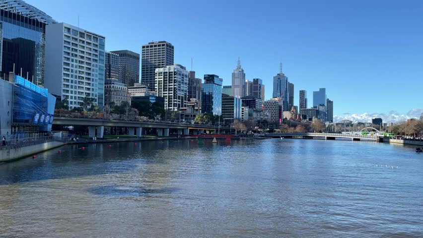 Slow motion video showcases the serene yarra river flowing through melbourne, highlighting the citys dynamic skyline against a clear blue sky, with birds gracefully flying in the distance and boats moving calmly on the water.