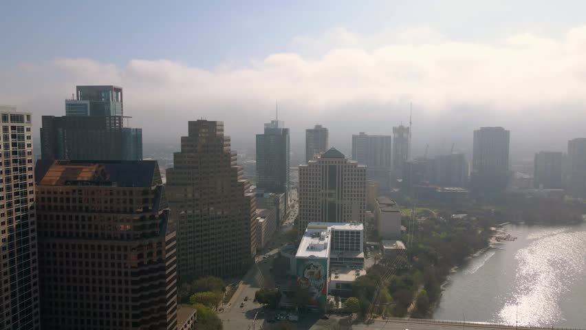 Dynamic drone shot moving across Austin cityscape during misty morning. View of high rise architecture, Congress Avenue Bridge, Lady Bird Lake, and urban development under soft sunlight.