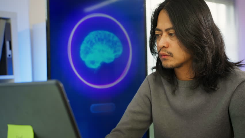 Focused Asian man typing on laptop while using artificial intelligence assistant tool in modern workspace. Generative AI technology, productivity tools, individual freelance work, Tech professionals.