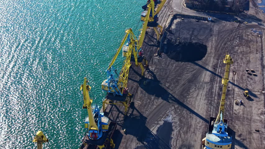 Cranes are positioned at the dock working to load cargo onto ships. The water is blue, and the site shows signs of industrial activity. The sun is shining on the cranes and dock area.