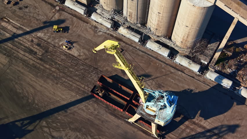 A crane operates on a construction site next to tall silos. Workers are present as the crane lifts a large container. Trucks are parked nearby on the ground.